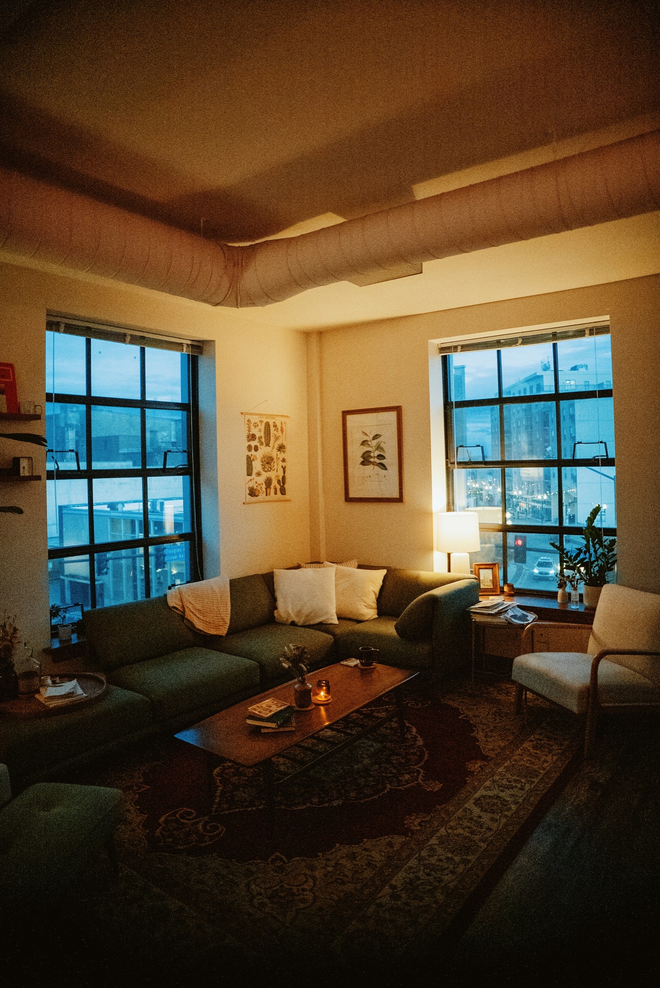 Lamp-lit sitting room at dusk with city windows